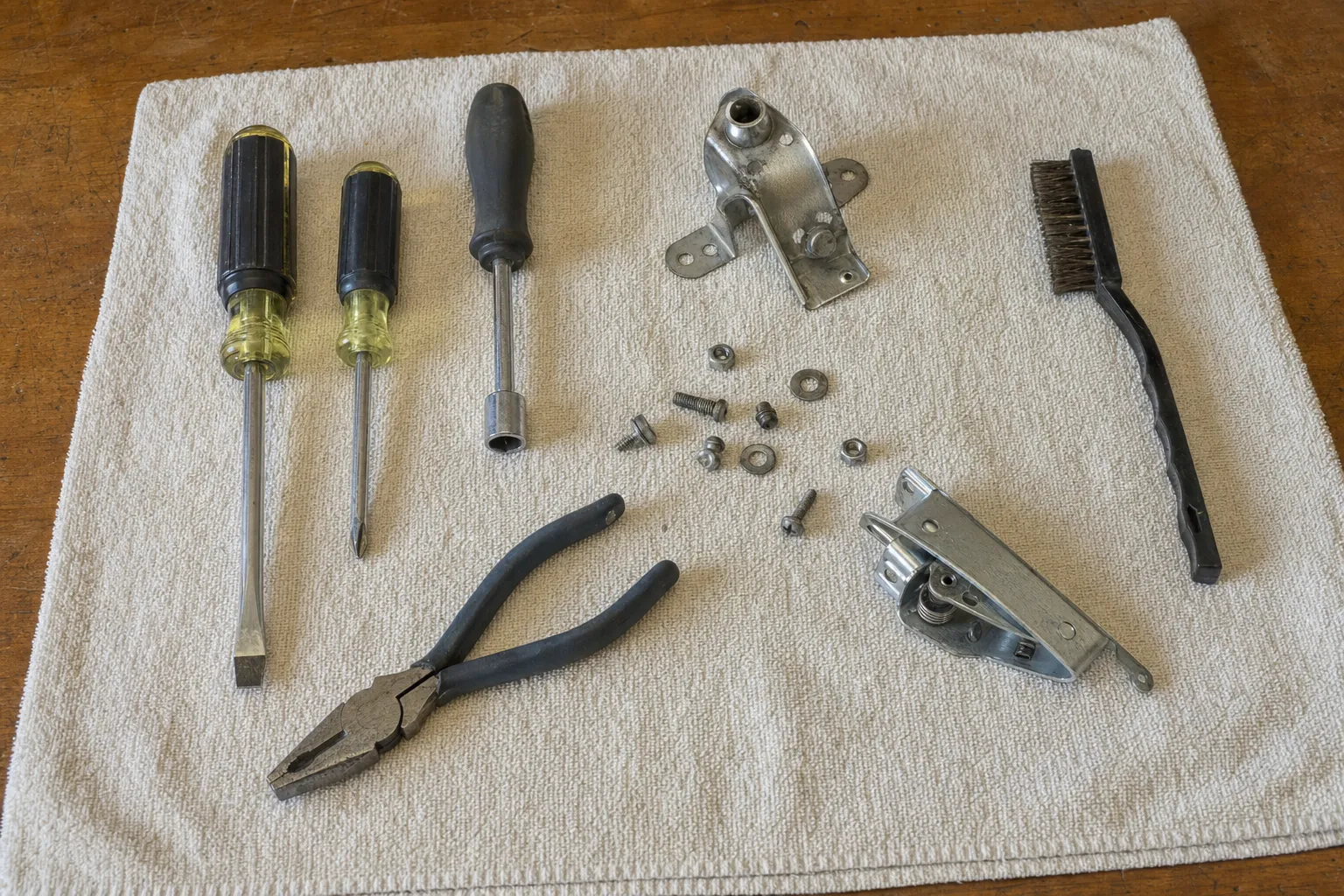 Screwdrivers, a nut driver, lineman pliers and a wire brush on a towel beside a bent stainless bracket, a sprung hinge assembly and loose screws, nuts and washers